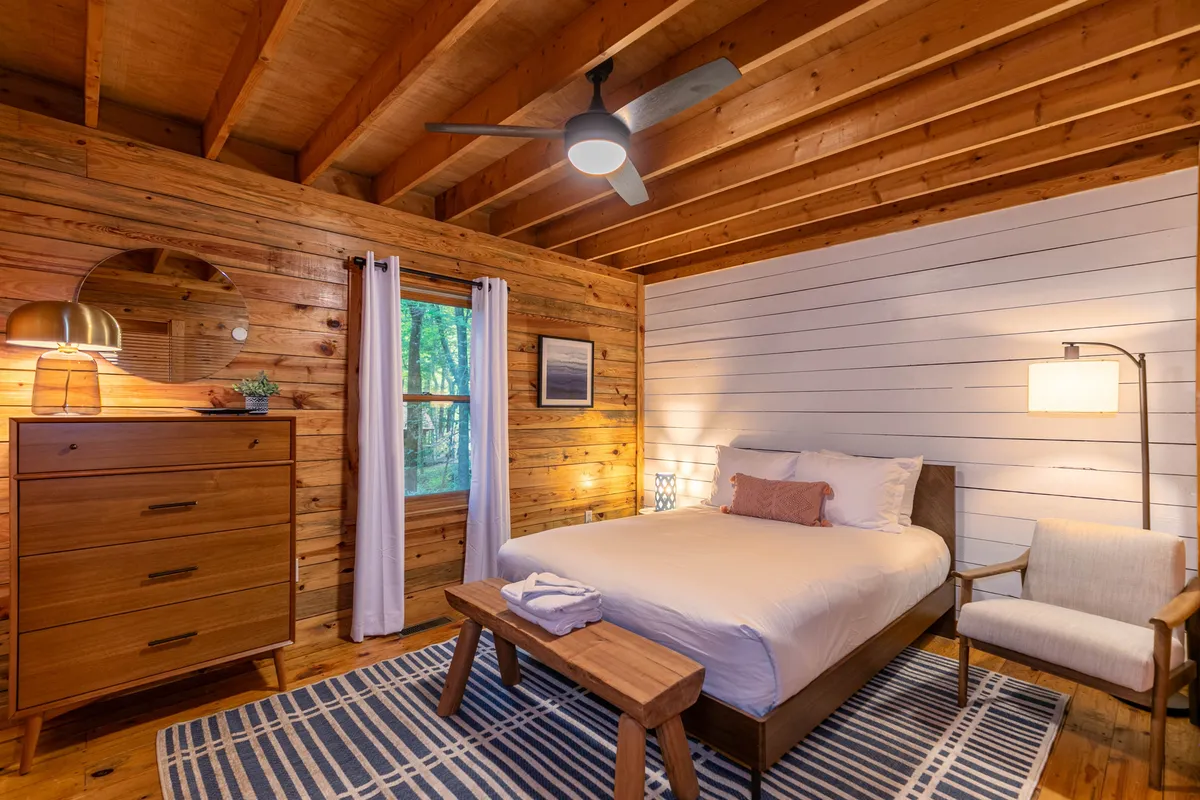 A serene bedroom featuring a bed with crisp white linens, a wooden dresser, a round mirror, an accent chair by the window, and soft natural light streaming in. The window reveals a tranquil view of the outdoors, enhancing the sense of privacy and relaxation.
