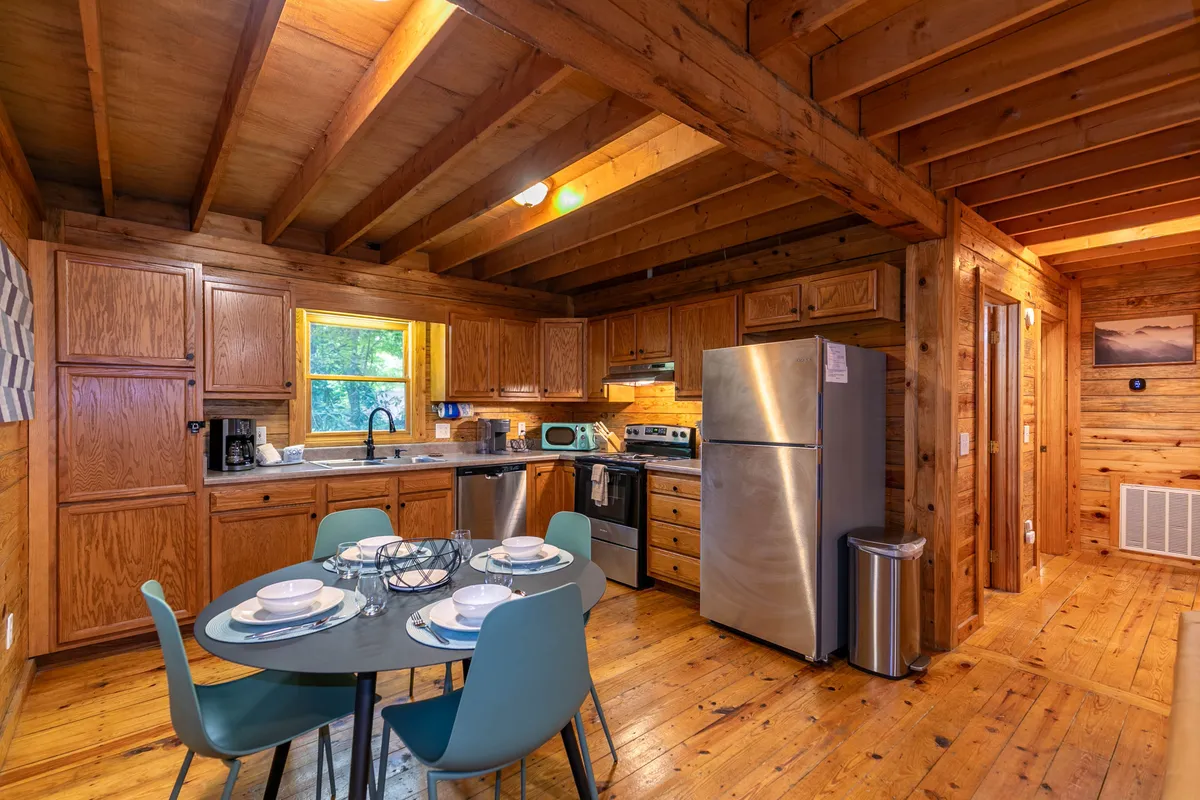 A sunlit dining area with a wooden table surrounded by chairs sits adjacent to a well-appointed kitchen. Natural light pours in through a window, illuminating modern appliances and rustic wooden beams overhead, creating a welcoming and stylish atmosphere.