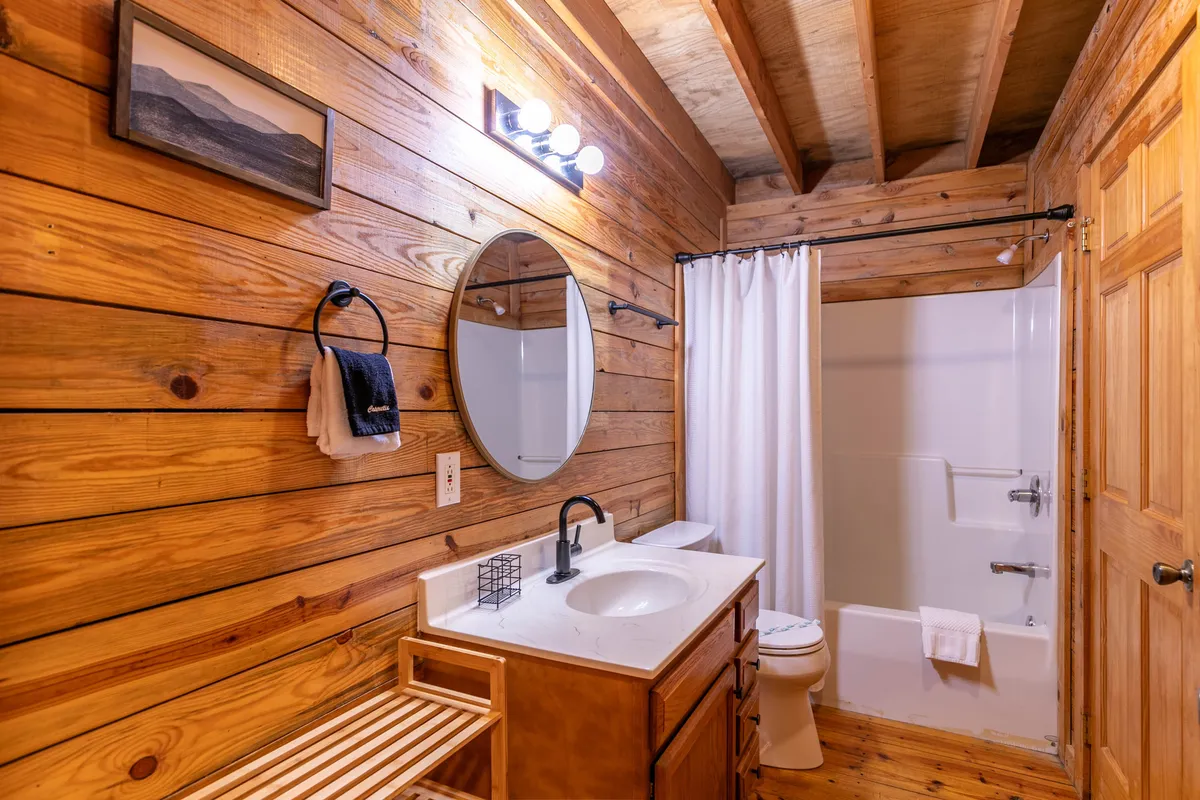 A warm, inviting en-suite bathroom with wooden walls, a round mirror above a stylish vanity, and a glimpse of the shower area. The overall vibe is tranquil and perfect for personal care or a relaxing soak.