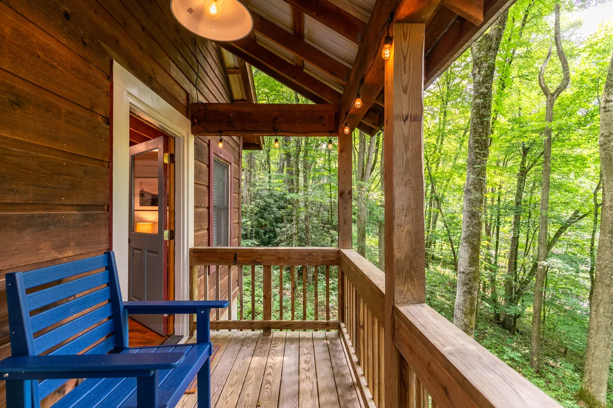 The image shows a cozy wooden porch with a blue chair, rustic railings, and a hanging light fixture, all overlooking a lush green forest. The serene setting invites guests to relax, unwind, and soak in the beauty of the surrounding nature.