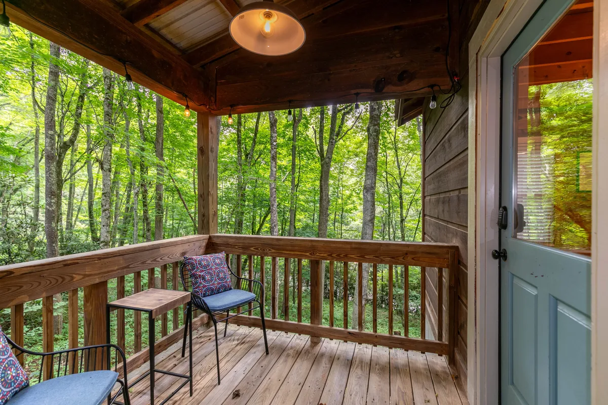 This image features a wooden deck with comfortable seating, a sturdy wooden railing, and an outdoor light fixture, all set against a backdrop of lush green trees. The angle looks out toward the forest, emphasizing the deck’s close connection to nature.