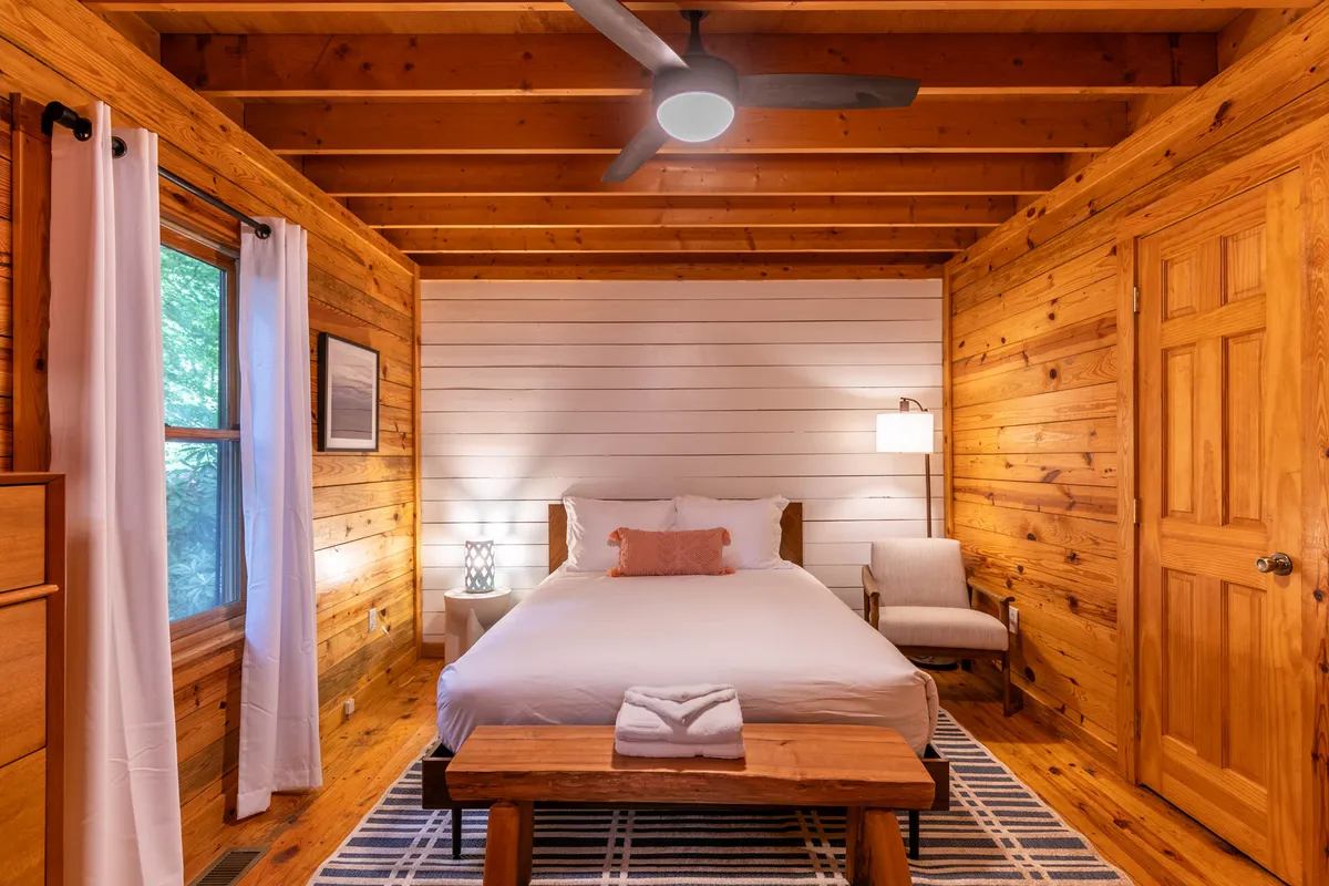 A serene bedroom featuring a bed draped in crisp white linens, a wooden bench at its foot, and warm wooden walls. Sunlight streams in, with a ceiling fan overhead, suggesting a peaceful, private retreat immersed in nature.