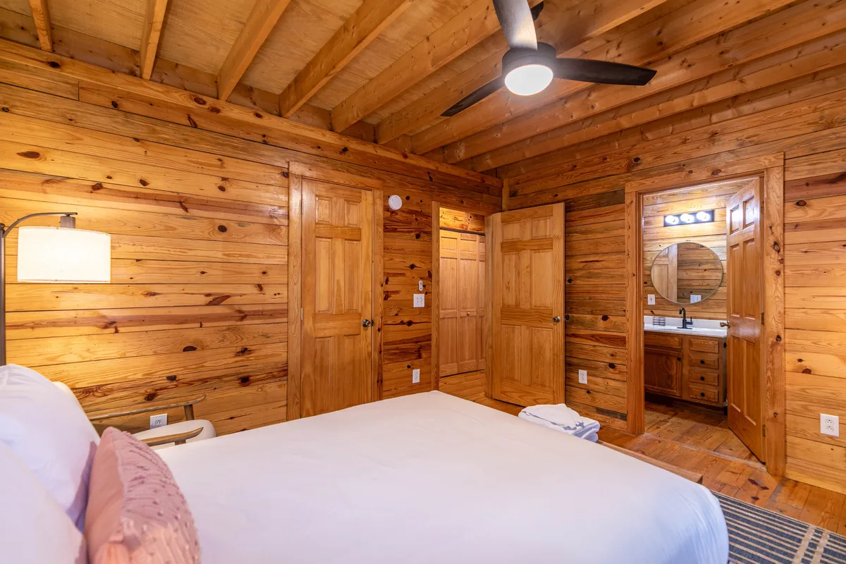 A cozy bedroom clad in warm wooden walls, featuring a comfortable bed with white linens, a bedside lamp, and a visible entrance to an en-suite bathroom. The ambiance is intimate, calm, and tailored for relaxation.