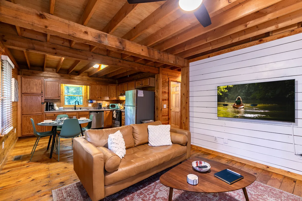 A perspective from the living area looking toward the dining space and open kitchen, featuring a plush leather sofa, a dining table set for sharing meals, and a TV displaying a serene nature scene. The environment is both warm and functional.
