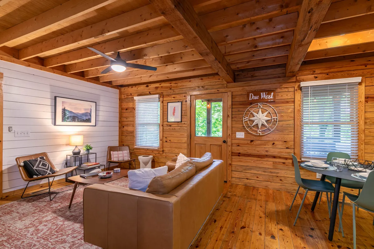A wide view of the main living area with a plush leather sofa, a wooden ceiling that enhances the rustic atmosphere, a dining table perfectly positioned for shared meals, tasteful wall art, and sunlight streaming through large windows. The inviting arrangement is ideal for gatherings—whether chatting, dining, or relaxing.