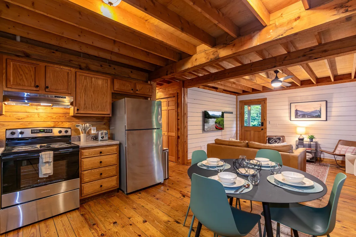 A cozy, open-plan scene featuring the kitchen with modern appliances, a wooden dining table ready for meals or conversation, a comfortable sofa, and artwork adorning the wall. The setup suggests activity, relaxation, and togetherness in one harmonious space.